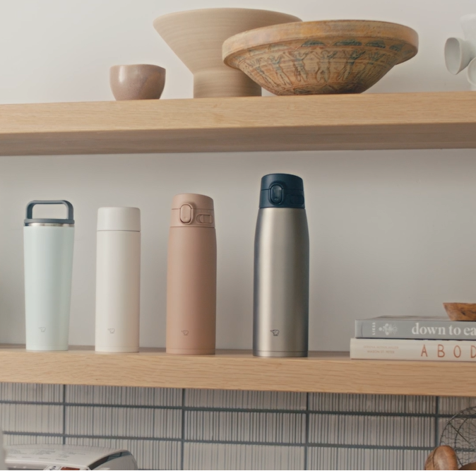 Set of four insulated travel mugs on a wooden shelf with decorative bowls and books in the background.