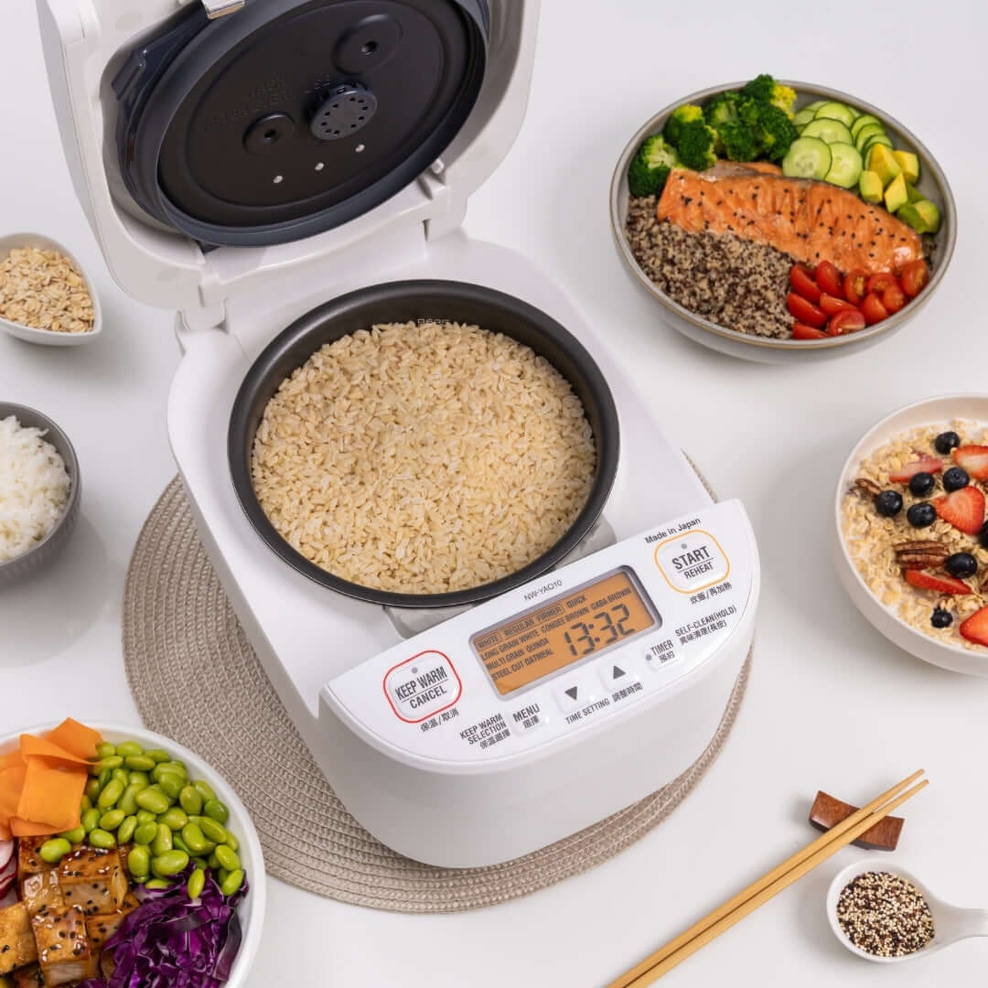 White rice cooker with brown rice on a white surface surrounded by various dishes and ingredients.