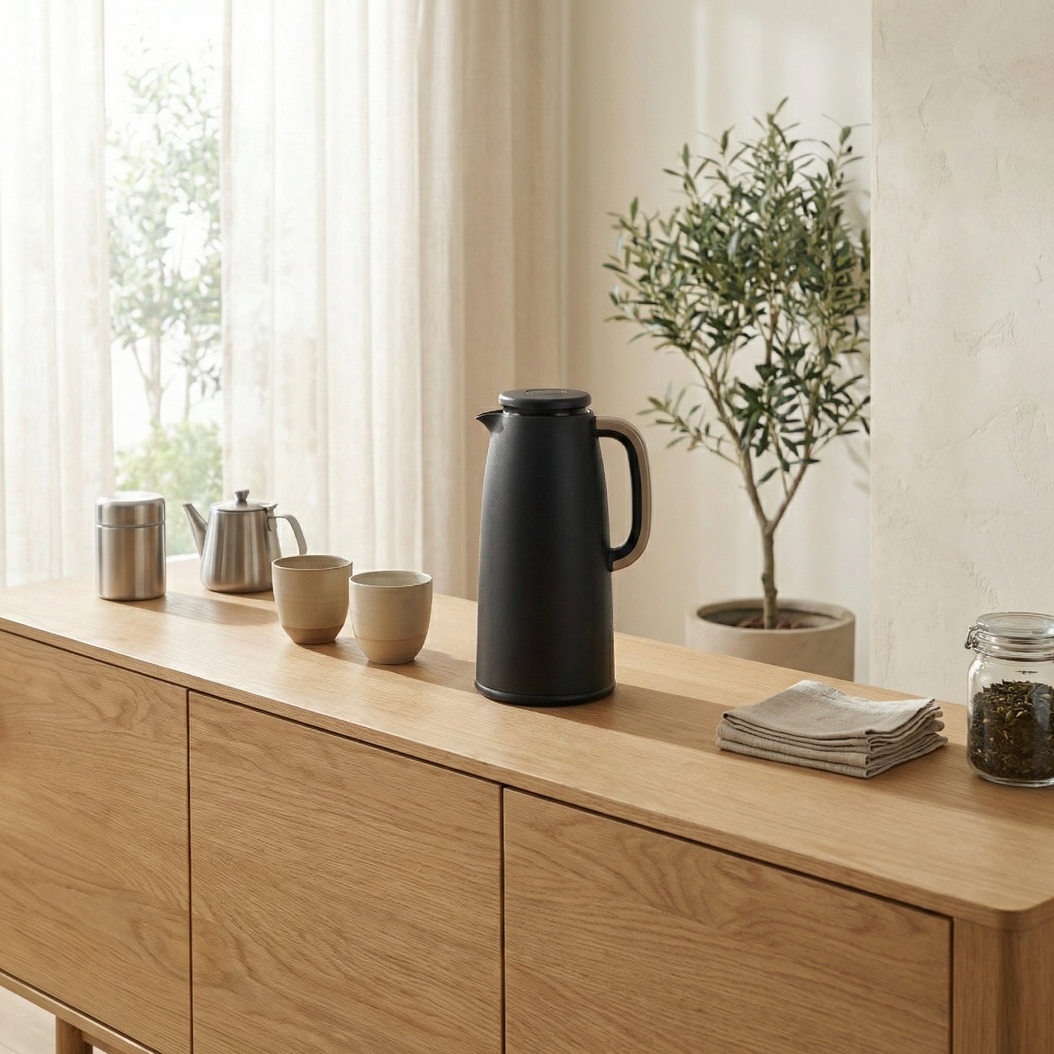 Black thermos on a wooden surface with cups and a plant in the background